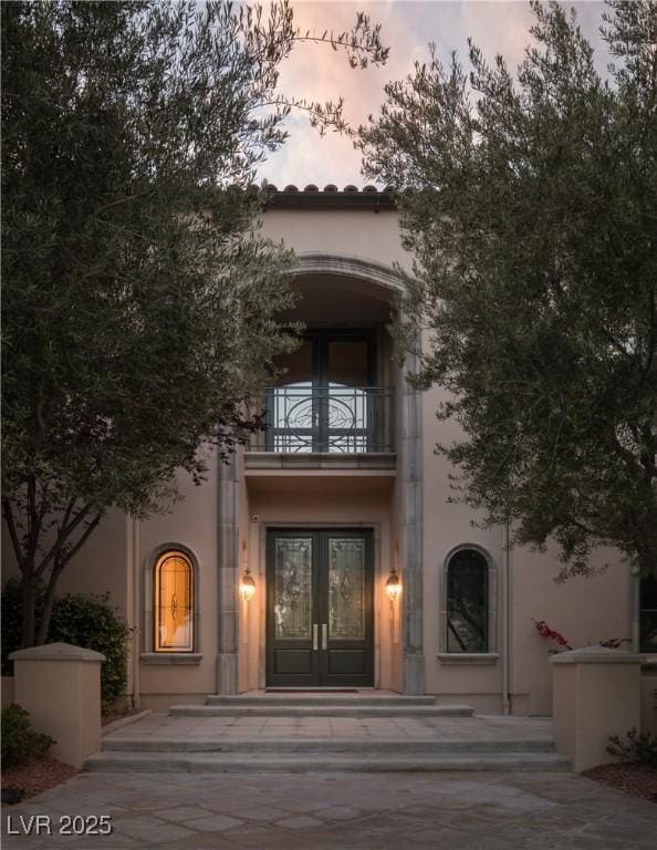 exterior entry at dusk with french doors, a balcony, and stucco siding