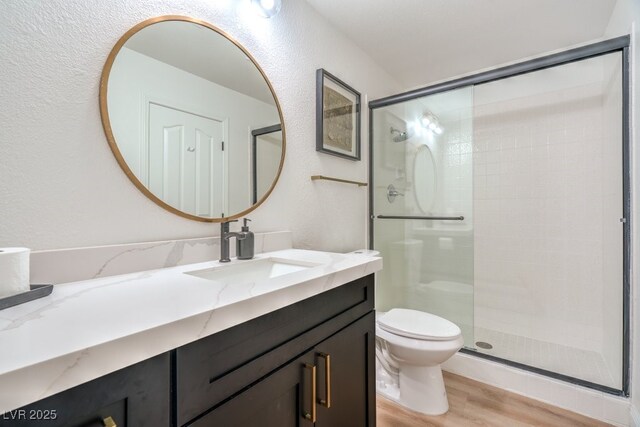 full bath with vanity, a textured wall, wood finished floors, toilet, and a stall shower