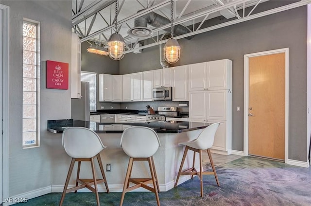 kitchen with dark countertops, appliances with stainless steel finishes, white cabinetry, a peninsula, and a towering ceiling