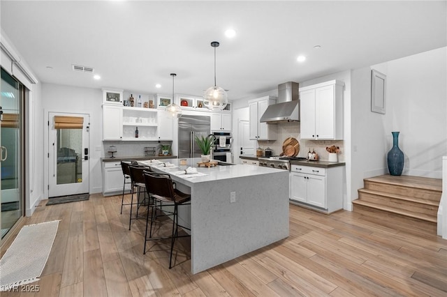 kitchen with a breakfast bar area, appliances with stainless steel finishes, tasteful backsplash, open shelves, and wall chimney exhaust hood