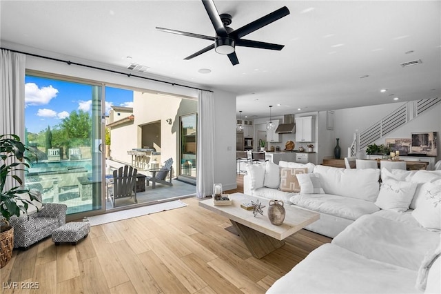 living room featuring visible vents, light wood finished floors, ceiling fan, and stairs
