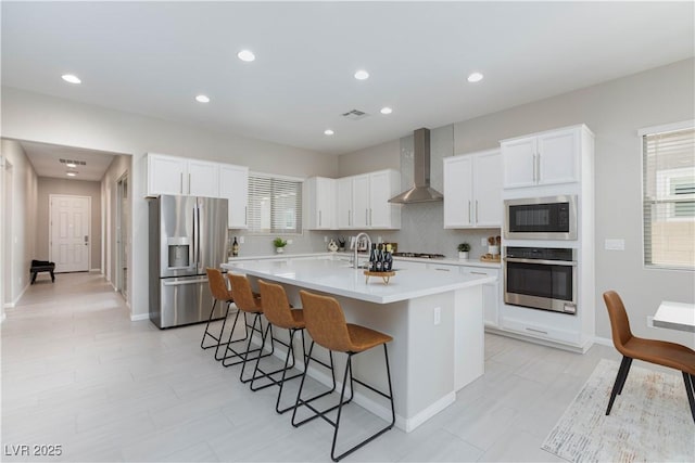 kitchen with wall chimney range hood, a kitchen island with sink, visible vents, appliances with stainless steel finishes, and a breakfast bar