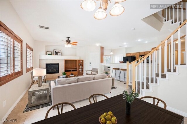 dining area with visible vents, a glass covered fireplace, baseboards, ceiling fan with notable chandelier, and light tile patterned floors