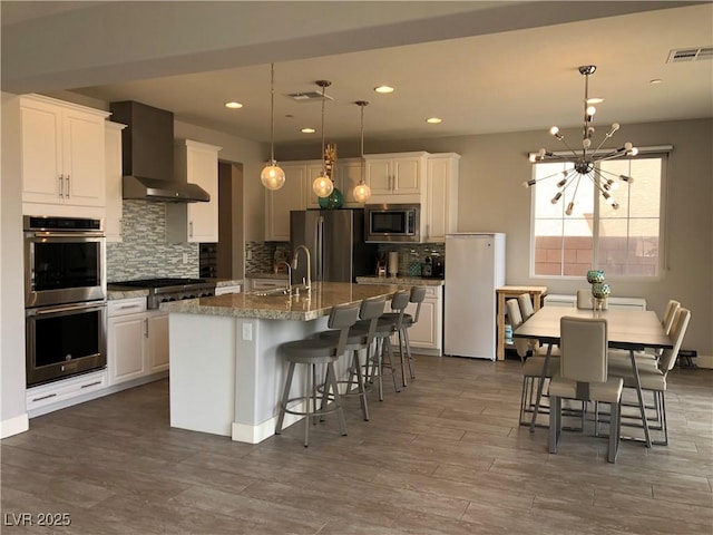 kitchen with wall chimney range hood, decorative backsplash, visible vents, a sink, and stainless steel appliances