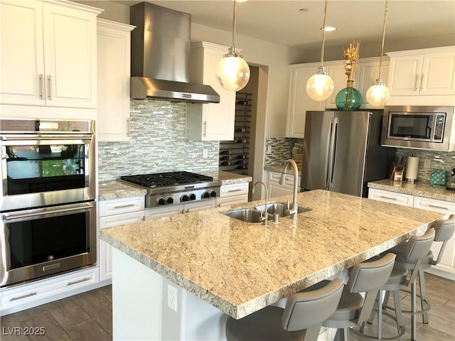 kitchen with wall chimney range hood, appliances with stainless steel finishes, a sink, an island with sink, and decorative backsplash