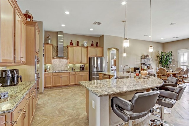 kitchen with light brown cabinets, wall chimney range hood, arched walkways, a sink, and stainless steel appliances