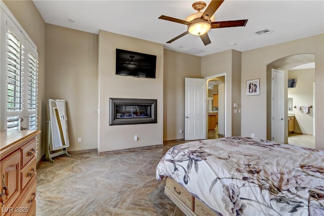 bedroom with ensuite bath, arched walkways, a glass covered fireplace, a ceiling fan, and visible vents