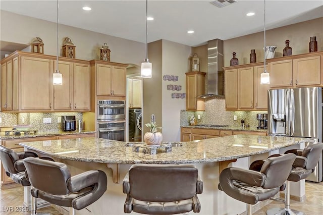 kitchen featuring wall chimney range hood, appliances with stainless steel finishes, light stone counters, a kitchen bar, and backsplash