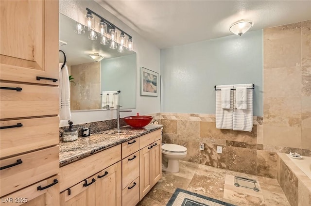 full bathroom with a relaxing tiled tub, vanity, toilet, stone finish floor, and tile walls