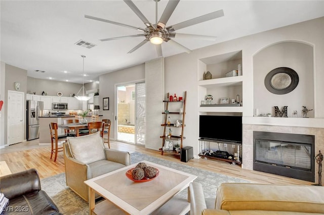 living area featuring visible vents, built in features, ceiling fan, a tile fireplace, and light wood-style flooring