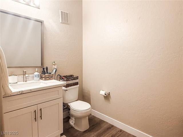 half bath featuring visible vents, vanity, a textured wall, wood finished floors, and toilet