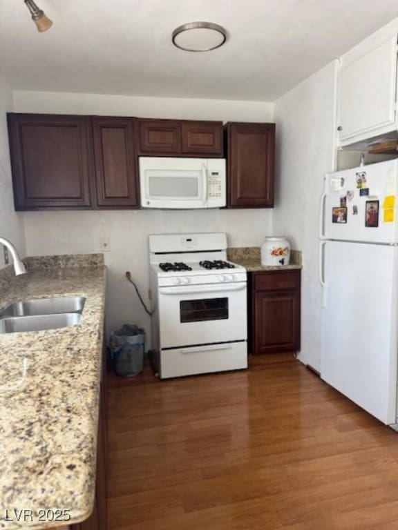 kitchen with dark brown cabinetry, light stone counters, a sink, white appliances, and wood finished floors