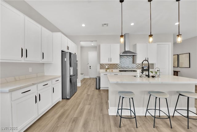 kitchen with stainless steel refrigerator with ice dispenser, light wood-style floors, a kitchen breakfast bar, and wall chimney range hood