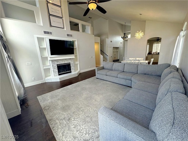 living area featuring a ceiling fan, dark wood-style flooring, built in shelves, stairs, and a stone fireplace