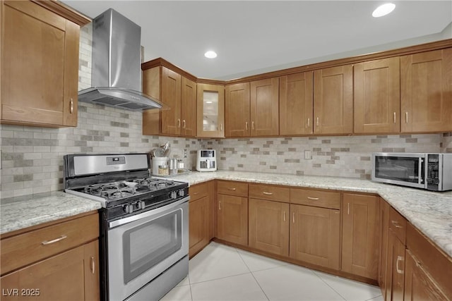 kitchen featuring light tile patterned flooring, appliances with stainless steel finishes, light stone counters, brown cabinetry, and wall chimney exhaust hood