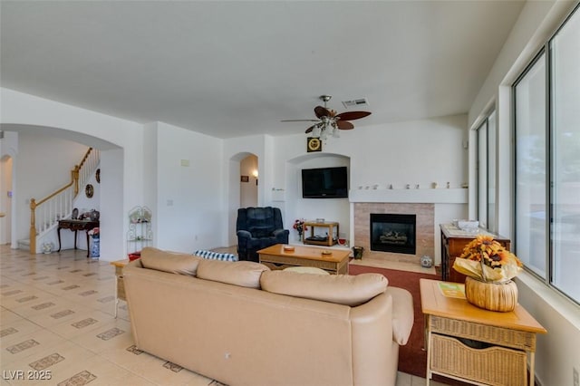 living area featuring arched walkways, a tiled fireplace, a ceiling fan, and stairway