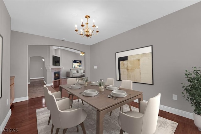 dining area with a warm lit fireplace, baseboards, ceiling fan with notable chandelier, arched walkways, and dark wood-style floors