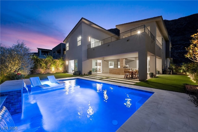 back of property at dusk with stucco siding, a balcony, a yard, an outdoor pool, and a patio area