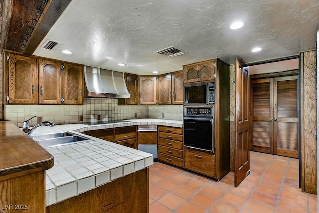 kitchen with a sink, tile counters, visible vents, black appliances, and wall chimney range hood