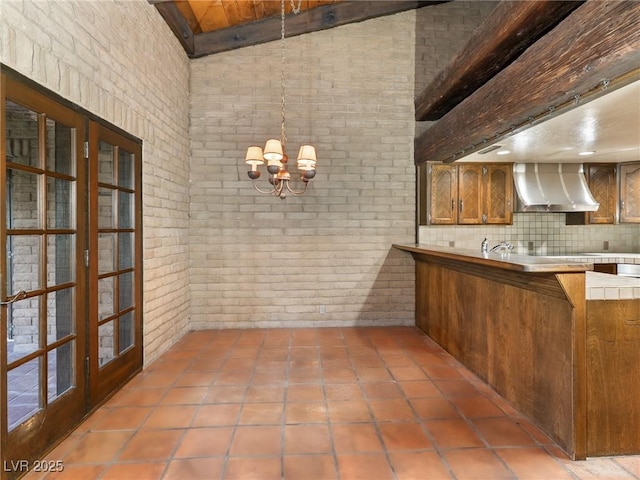 kitchen featuring a notable chandelier, brick wall, decorative backsplash, wall chimney exhaust hood, and beamed ceiling