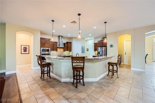 kitchen featuring stainless steel appliances, wall chimney exhaust hood, arched walkways, a kitchen bar, and light stone counters