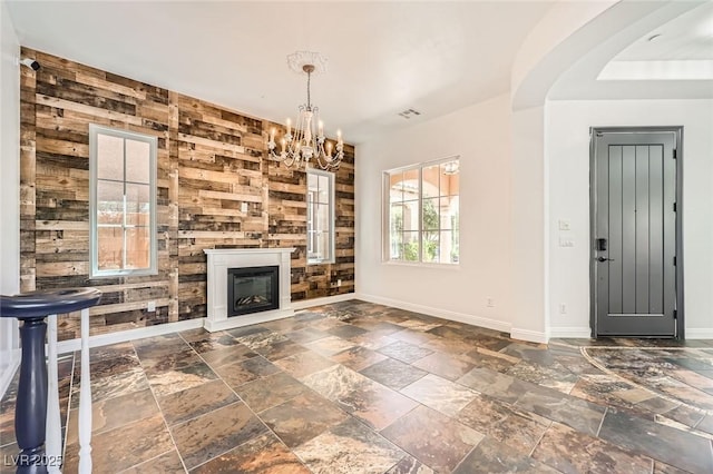 unfurnished living room featuring wood walls, stone tile flooring, a notable chandelier, baseboards, and a glass covered fireplace