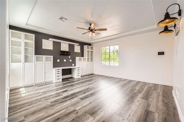 unfurnished living room with built in desk, wood finished floors, a tray ceiling, baseboards, and ceiling fan
