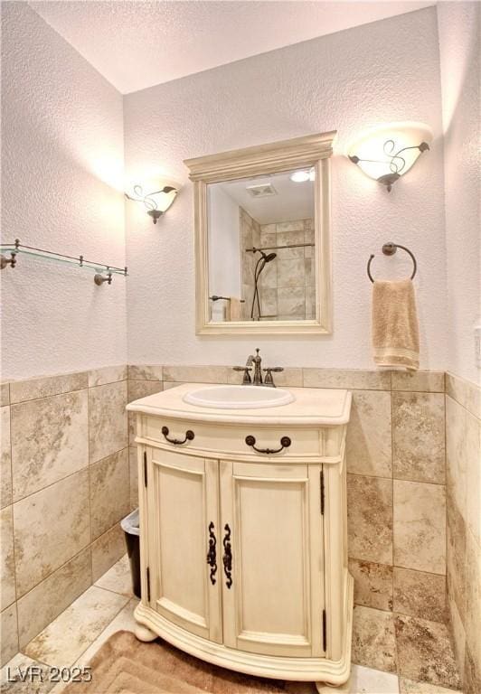 bathroom with tile walls, vanity, a textured wall, a tile shower, and a wainscoted wall