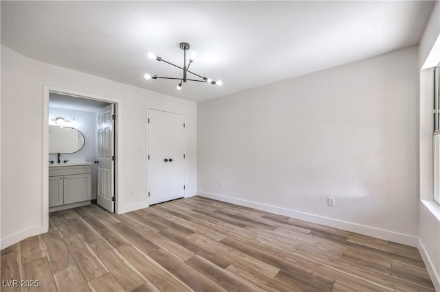 unfurnished bedroom featuring a notable chandelier, baseboards, ensuite bath, a sink, and light wood-style floors