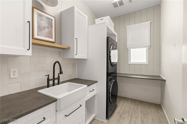 kitchen with stacked washing machine and dryer, dark countertops, a sink, white cabinetry, and light wood-type flooring