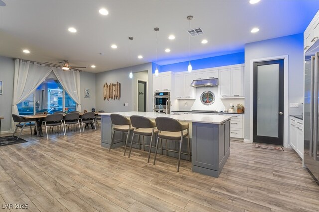 5865 Alice Landing Ct, Las Vegas, NV 89149 - kitchen with under cabinet range hood, stovetop, a kitchen island with sink, light wood-style flooring, and white cabinetry