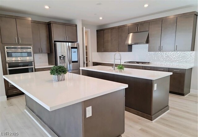 kitchen featuring a kitchen island with sink, under cabinet range hood, stainless steel appliances, light wood-style floors, and dark brown cabinetry