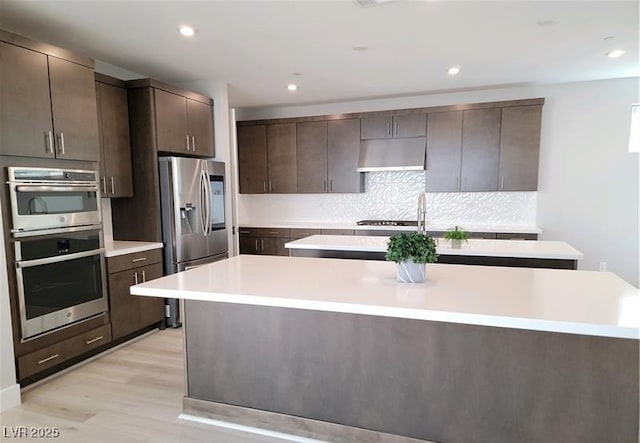 kitchen with stainless steel appliances, an island with sink, dark brown cabinets, light countertops, and recessed lighting