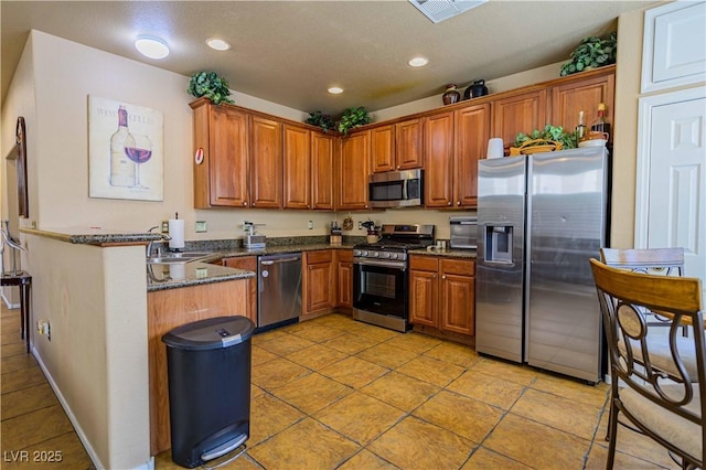 kitchen with stainless steel appliances, light tile patterned floors, brown cabinets, a sink, and dark stone countertops