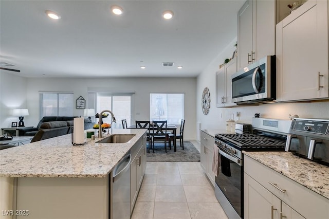 kitchen with stainless steel appliances, a sink, light colored carpet, a center island with sink, and light stone counters