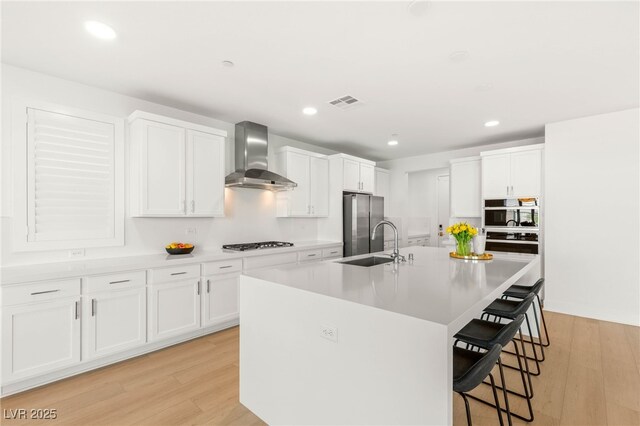 kitchen featuring wall chimney range hood, appliances with stainless steel finishes, a kitchen bar, light wood-style floors, and recessed lighting