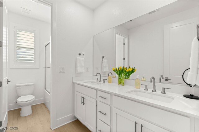 bathroom featuring toilet, wood finished floors, double vanity, baseboards, and enclosed tub / shower combo