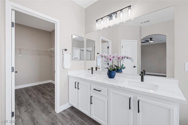 full bath featuring ceiling fan, wood finished floors, double vanity, a walk in closet, and baseboards
