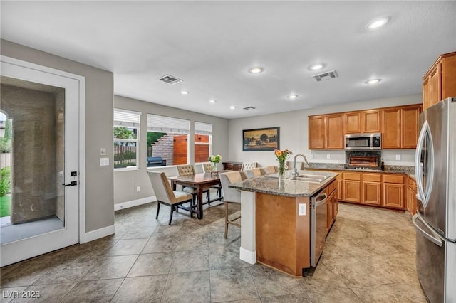 kitchen featuring stainless steel appliances, a sink, stone counters, a kitchen island with sink, and brown cabinets