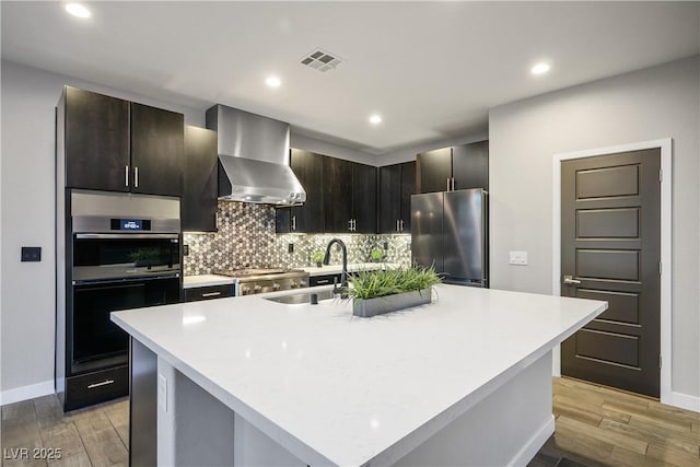 kitchen with stainless steel refrigerator, wall chimney range hood, a sink, double oven, and light wood-style flooring