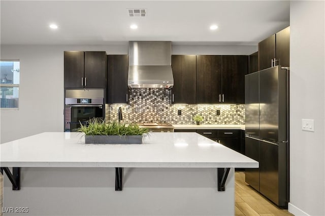 kitchen with appliances with stainless steel finishes, wall chimney range hood, decorative backsplash, light countertops, and a center island