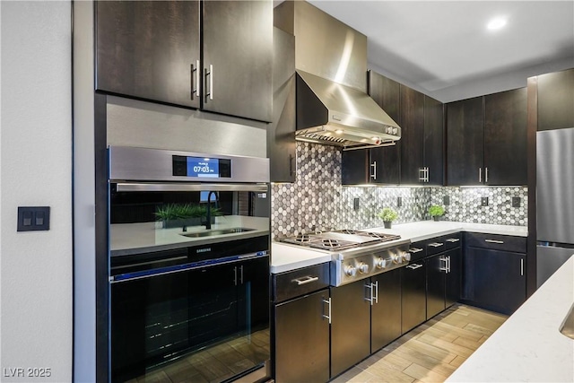 kitchen featuring appliances with stainless steel finishes, light countertops, light wood-style flooring, wall chimney exhaust hood, and tasteful backsplash