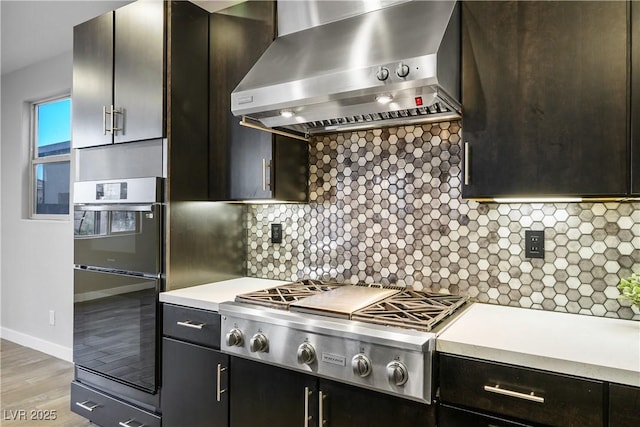 kitchen featuring wall chimney range hood, stainless steel gas stovetop, light countertops, multiple ovens, and tasteful backsplash