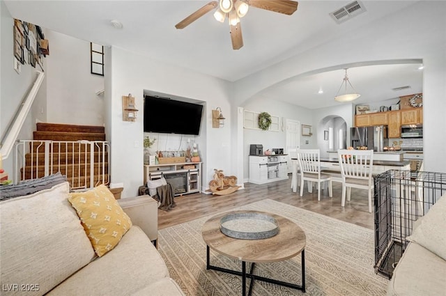 living area featuring arched walkways, light wood-type flooring, ceiling fan, stairs, and a fireplace