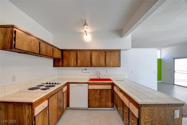 kitchen featuring a sink, a peninsula, white appliances, tile counters, and brown cabinets