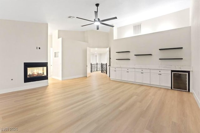 unfurnished living room featuring ceiling fan, beverage cooler, light wood-style floors, baseboards, and a towering ceiling