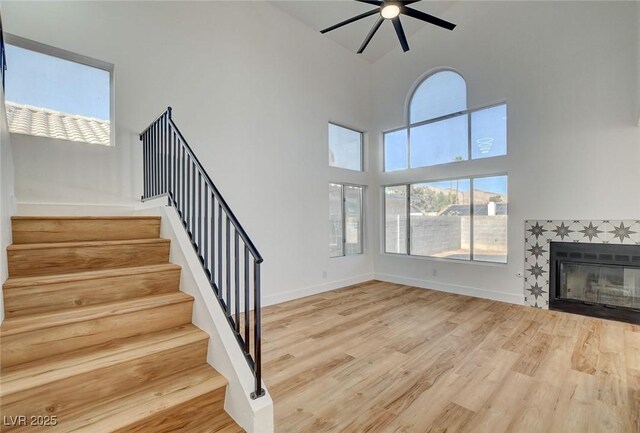 unfurnished living room with ceiling fan, wood finished floors, a towering ceiling, stairs, and a tile fireplace