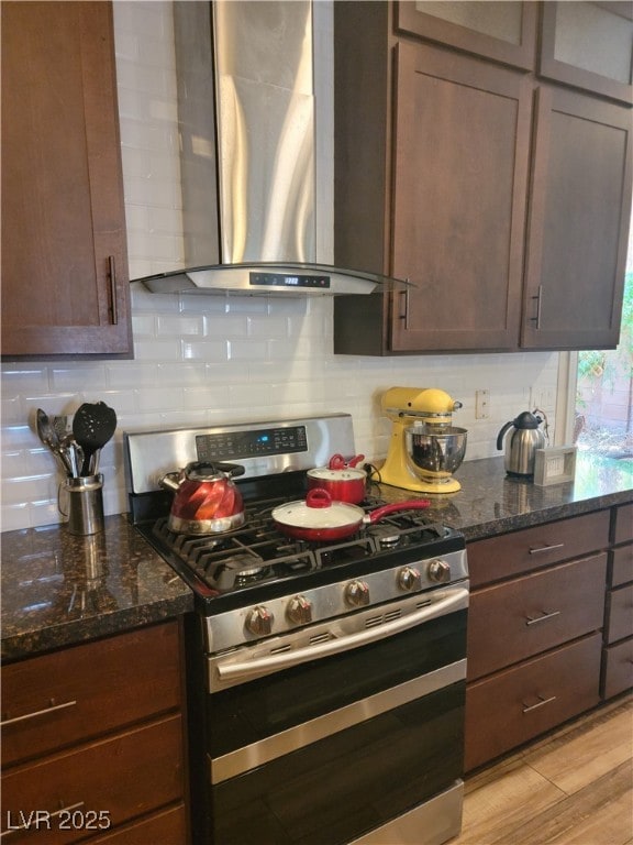 kitchen featuring wall chimney exhaust hood, stainless steel range with gas cooktop, decorative backsplash, dark stone counters, and dark brown cabinetry