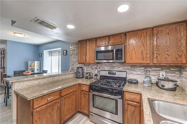 6408 Deadwood Rd, Las Vegas, NV 89108 - kitchen with stainless steel appliances, a peninsula, brown cabinets, a textured ceiling, and backsplash