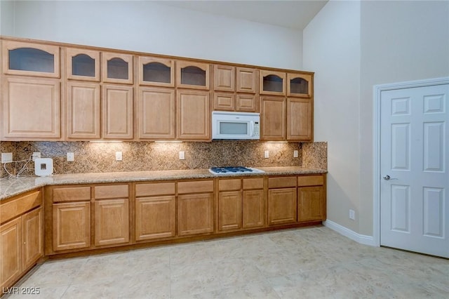 kitchen with white appliances, backsplash, glass insert cabinets, light stone counters, and baseboards